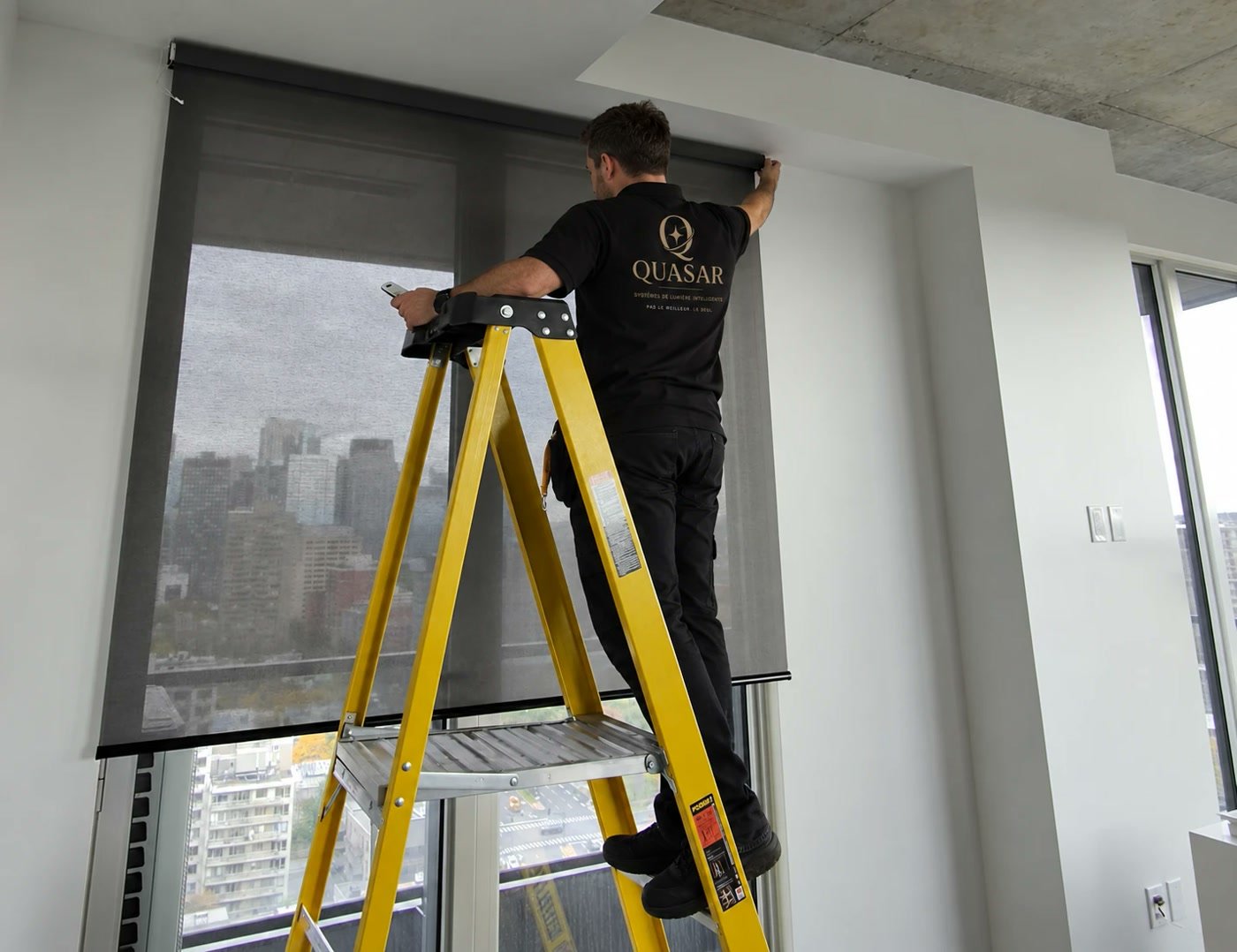 Un installateur Quasar sur un escabeau qui pose une toile enrouleuse en tête de fenêtre.